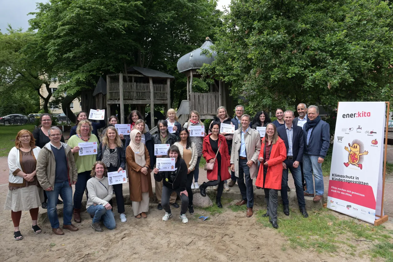 Gruppenfoto mit 20 P&auml;dagog*innen, die Pr&auml;mien in der Handhalten, rechts am Bild enerkita-Rollup und Bi&ouml;dungssenatorin Sascha Aulepp